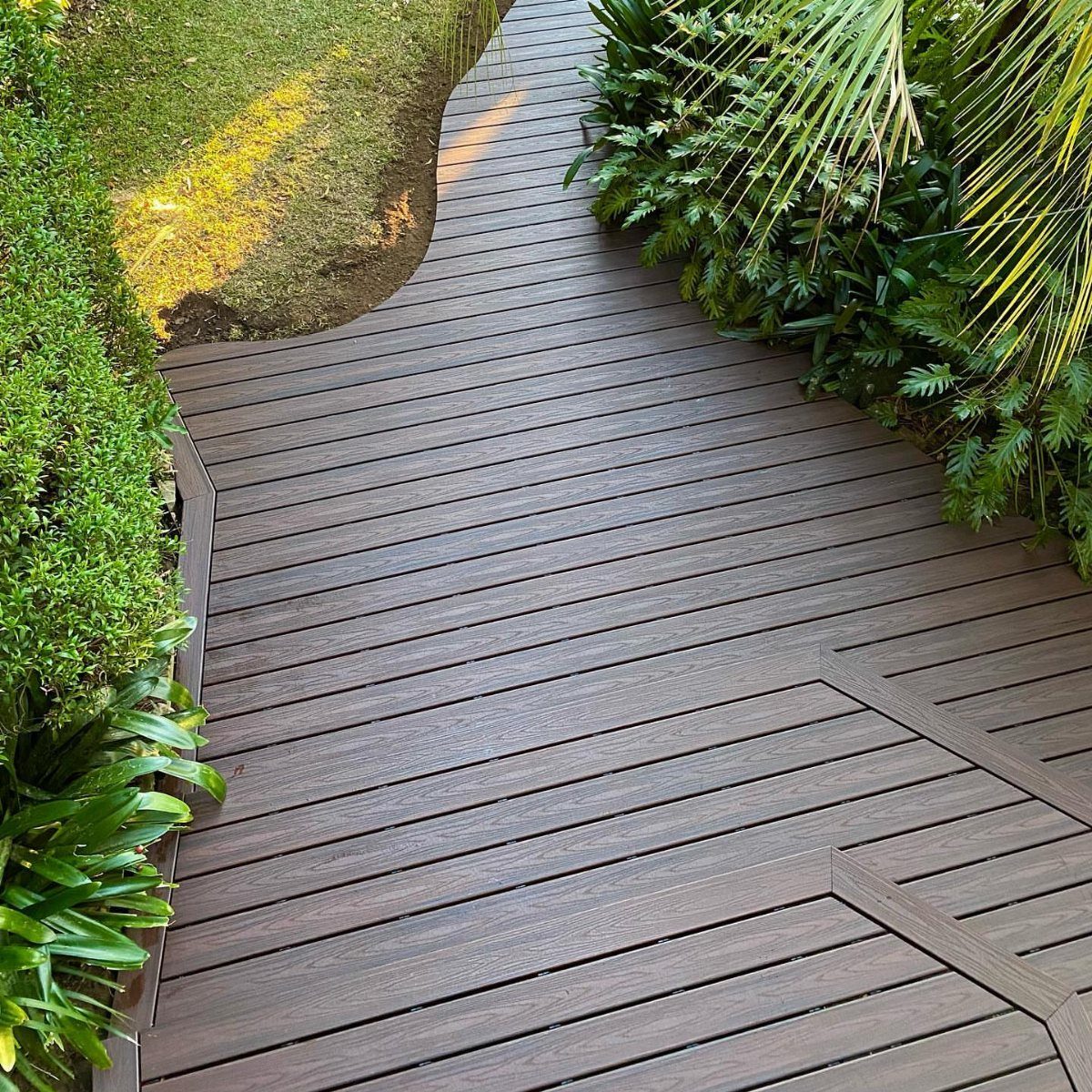 11 Around Deck Landscaping Ideas with Photos and Tips 11 A curved dark wood deck surrounded by lush green tropical plants and manicured garden shrubbery.