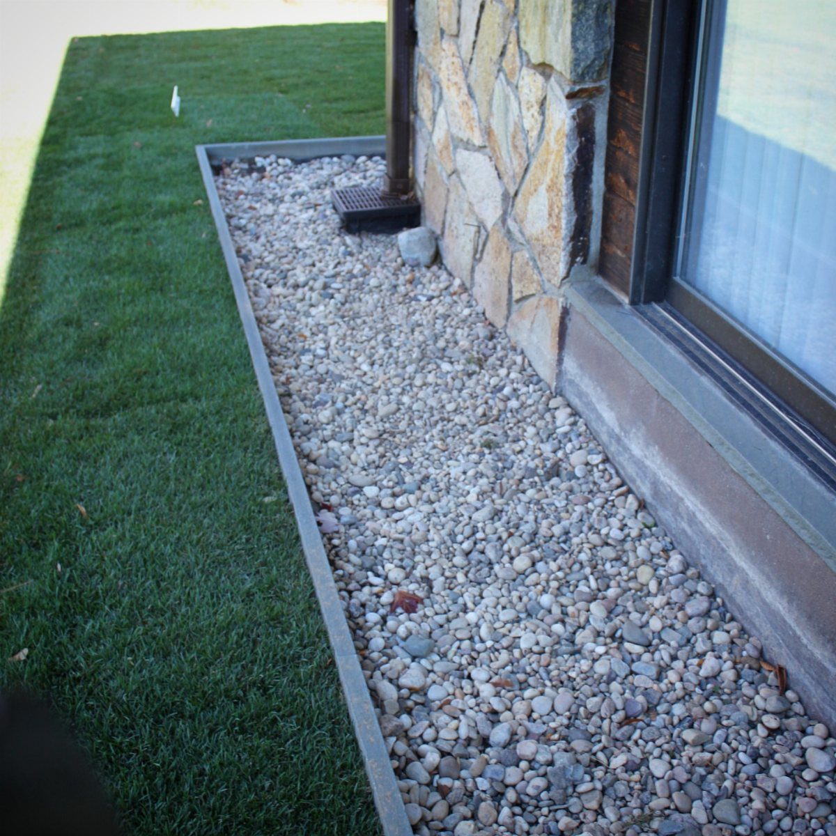 11 Around House Landscaping Ideas for Foundation to Fence 6 A gravel strip with grey stones runs along a stone house foundation next to green grass.