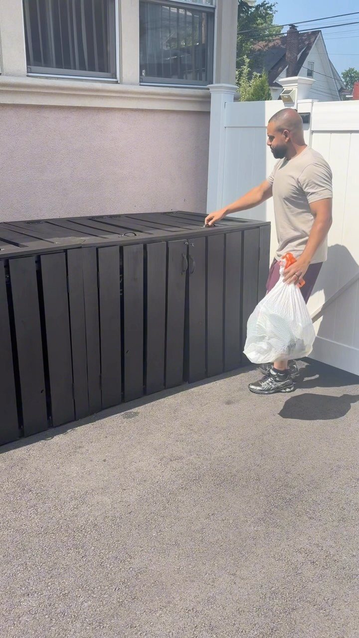 11 Around House Landscaping Ideas for Foundation to Fence 9 A man standing next to a black wooden trash bin enclosure on an asphalt driveway.