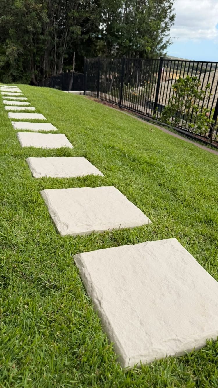 11 Around House Landscaping Ideas for Foundation to Fence 7 Large rectangular stone pavers create a stepping stone path through a green grass lawn next to a fence.