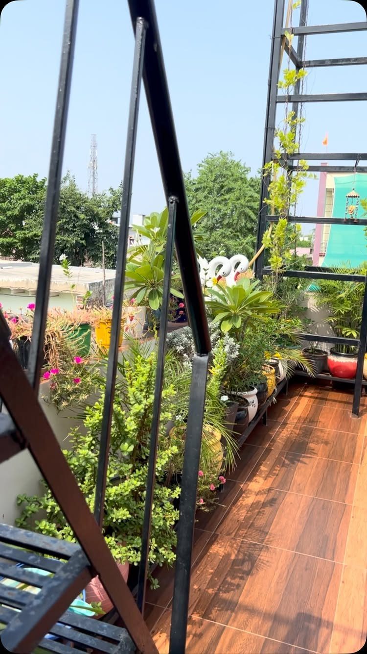 Potted green plants and flowers arranged along a narrow balcony with black metal railings and wooden tiles.