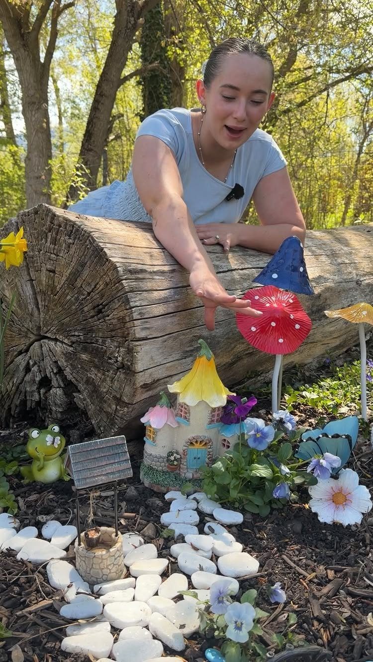 A young woman points at a whimsical miniature fairy garden with a small house and mushrooms.