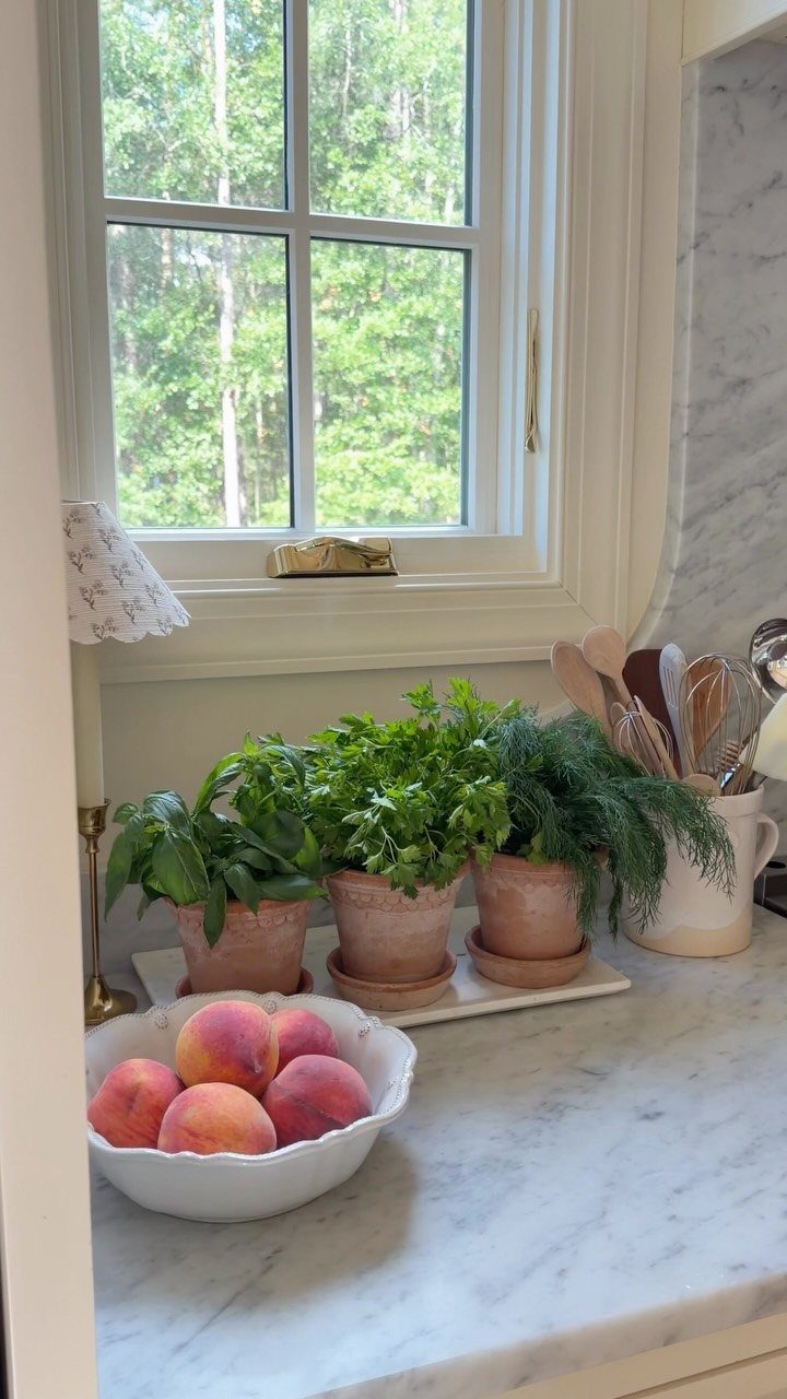 Three terracotta pots with green herbs sitting on a marble countertop by a window.