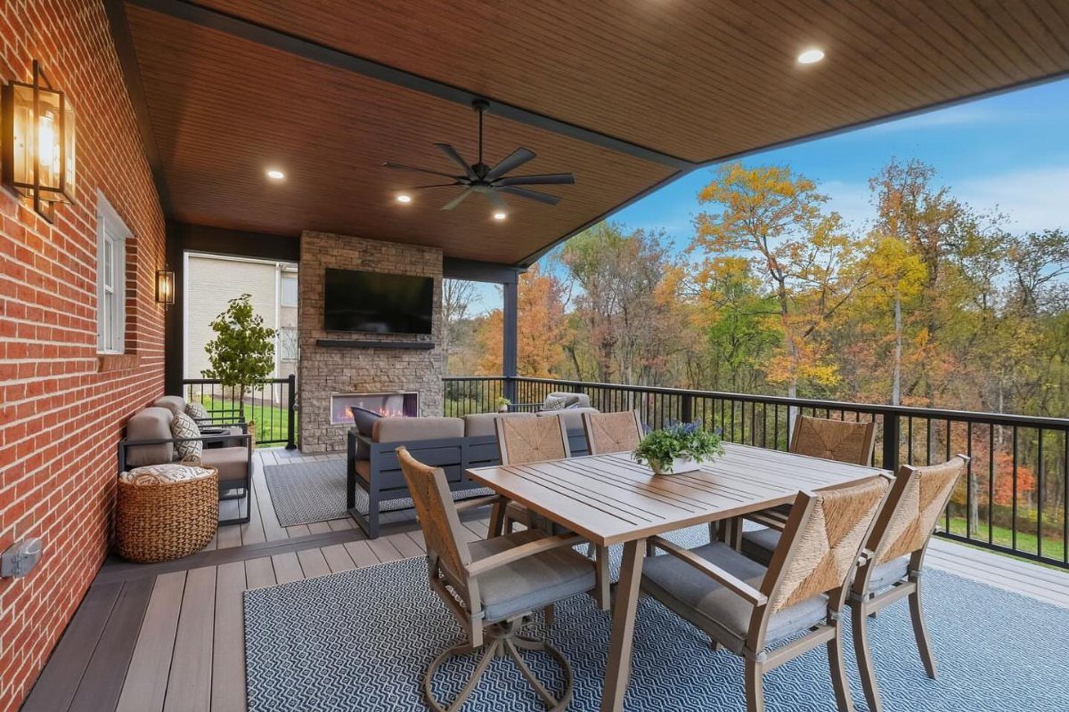 21 Backyard Deck Ideas For Any Size Space 9 A covered outdoor deck featuring a dining set, lounge seating, and a stone fireplace overlooking autumn trees.