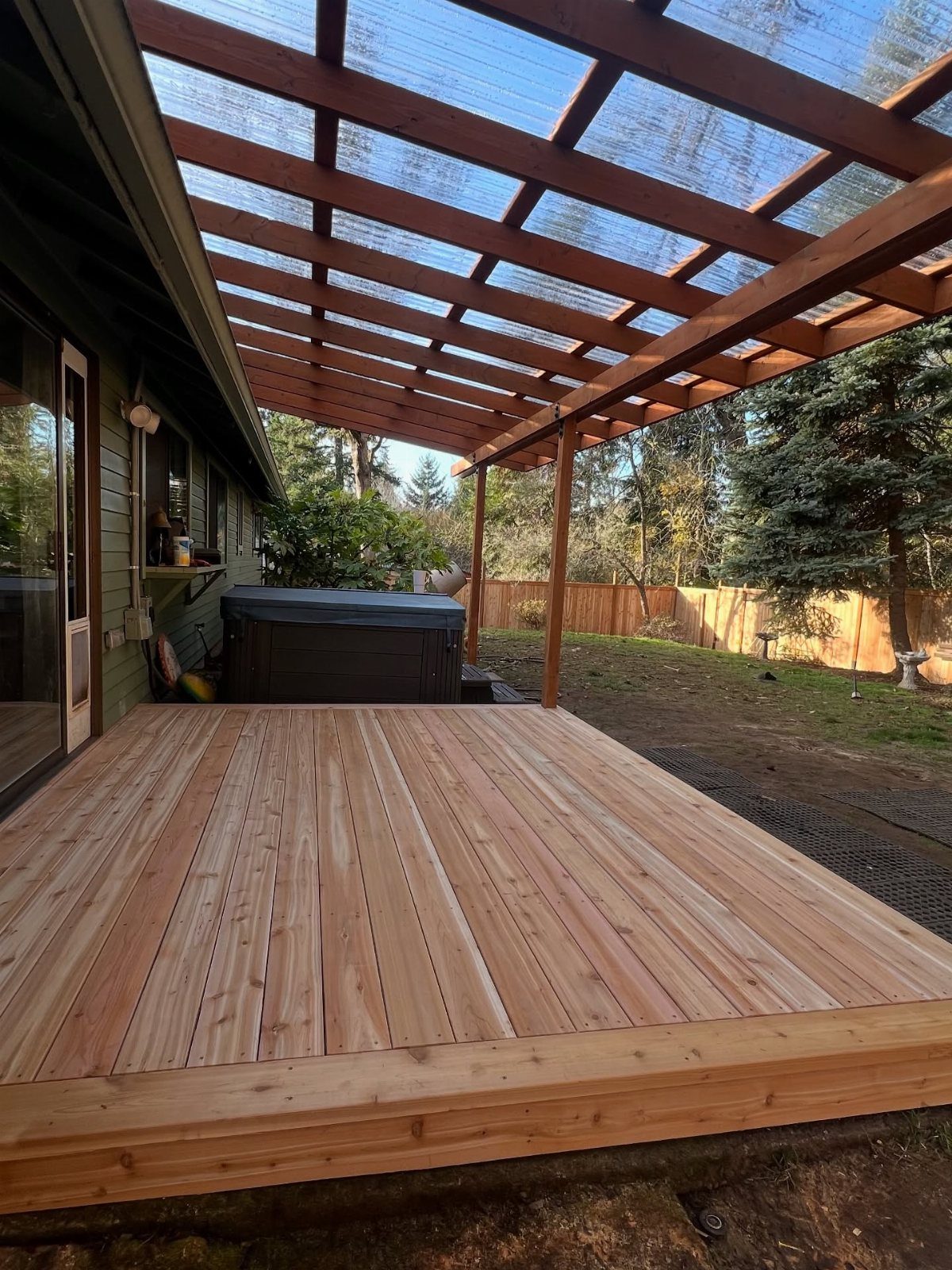 21 Backyard Deck Ideas For Any Size Space 1 A wooden deck with a translucent roofed pergola attached to a house near trees.