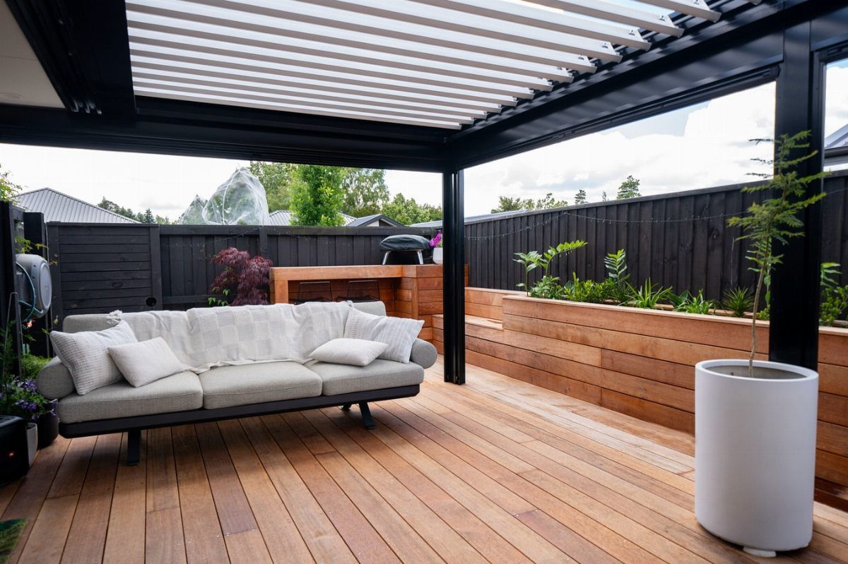 21 Backyard Deck Ideas For Any Size Space 4 Modern wooden deck with a grey sofa, black pergola, and built-in wooden planter boxes.