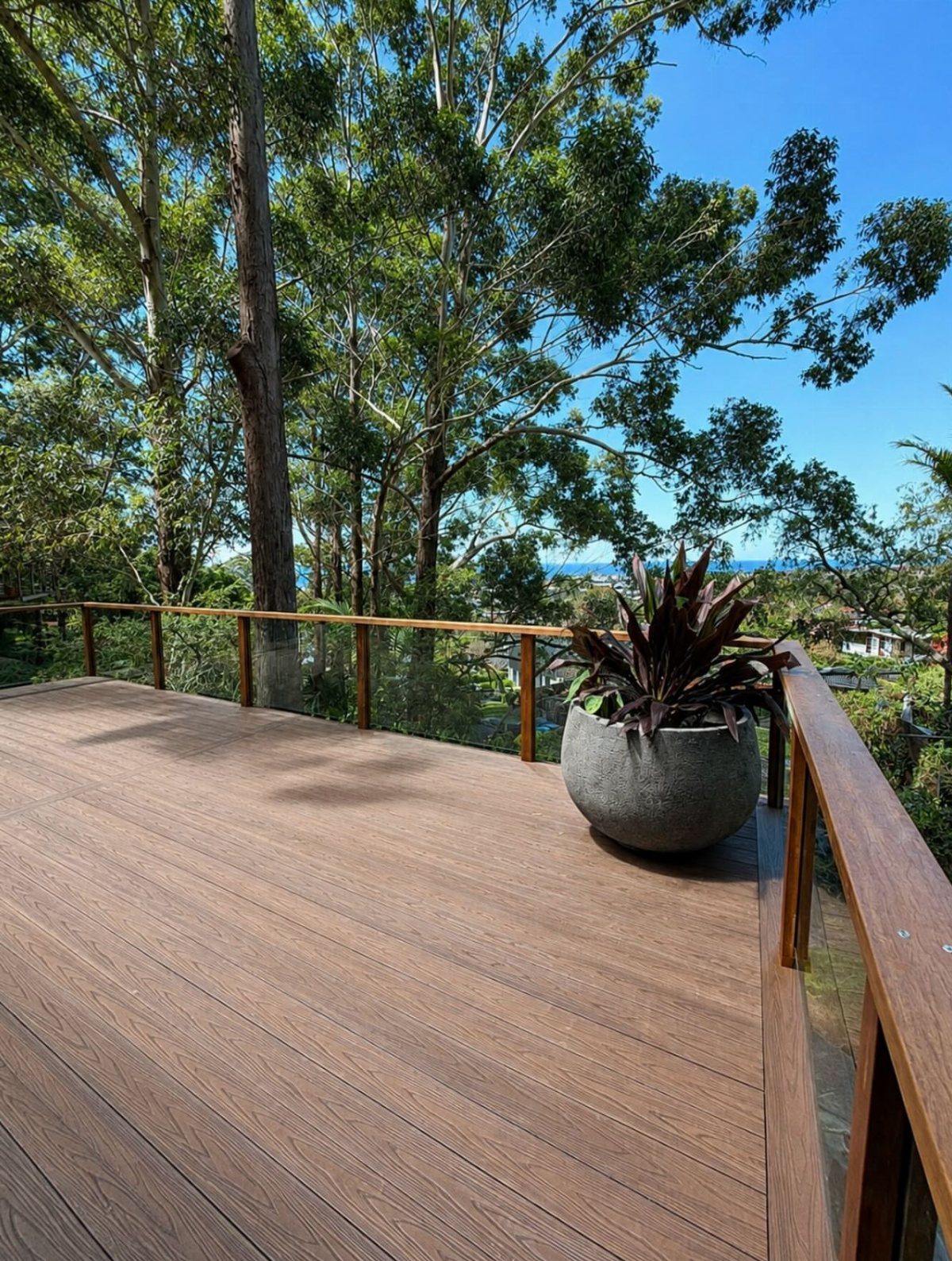 21 Backyard Deck Ideas For Any Size Space 8 Wooden deck with glass railings overlooking green trees and the blue ocean under a clear sky.