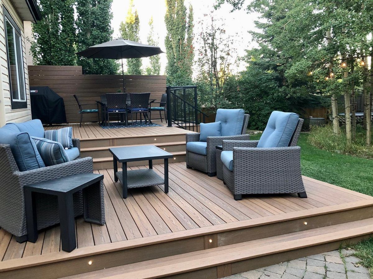 21 Backyard Deck Ideas For Any Size Space 11 Multi-level wooden deck with grey wicker furniture and integrated lighting in a backyard setting.