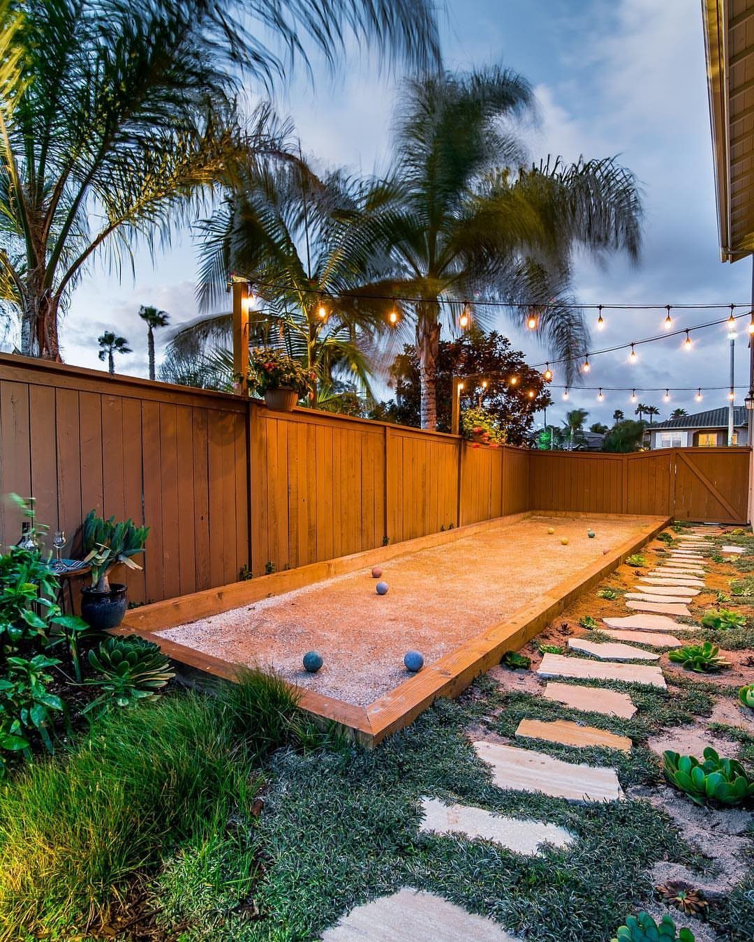 A rectangular sand bocce ball court with colorful balls sits in a landscaped backyard.