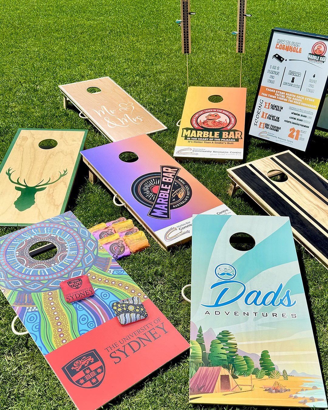 Multiple colorful custom-designed cornhole boards scattered across a green grass lawn.