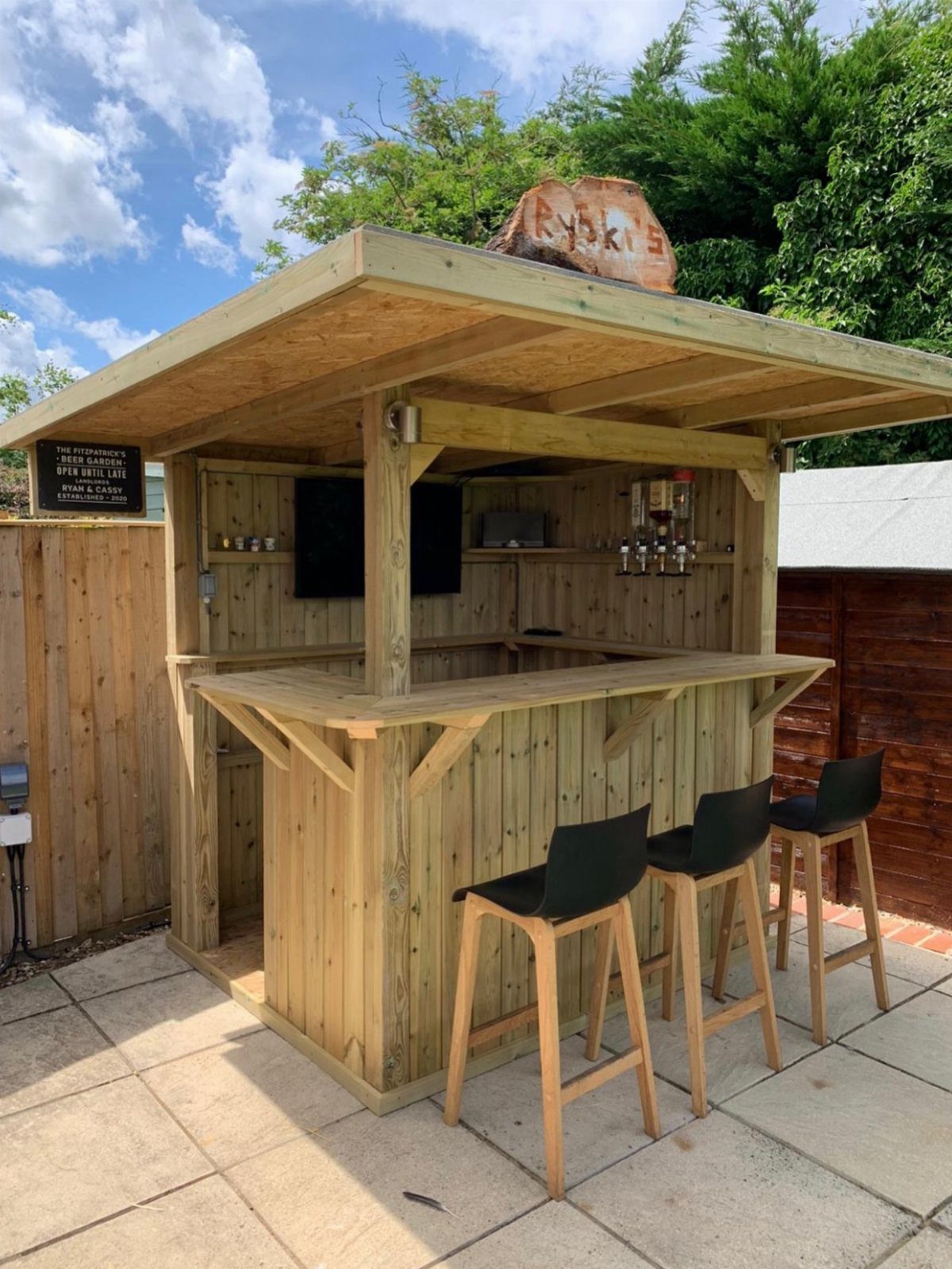 A wooden outdoor bar structure with three black stools on a paved patio.