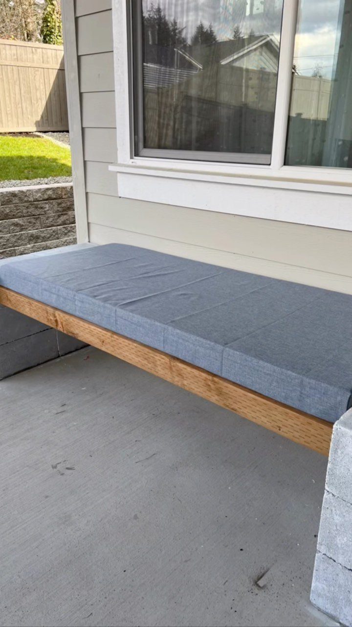A long wooden outdoor bench with a grey cushion mounted against a house wall.