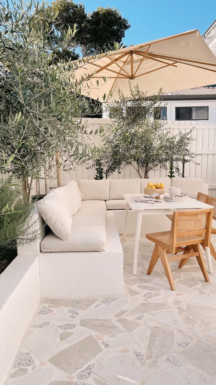 White built-in outdoor sectional sofa with cream cushions on a stone patio under a tan umbrella.