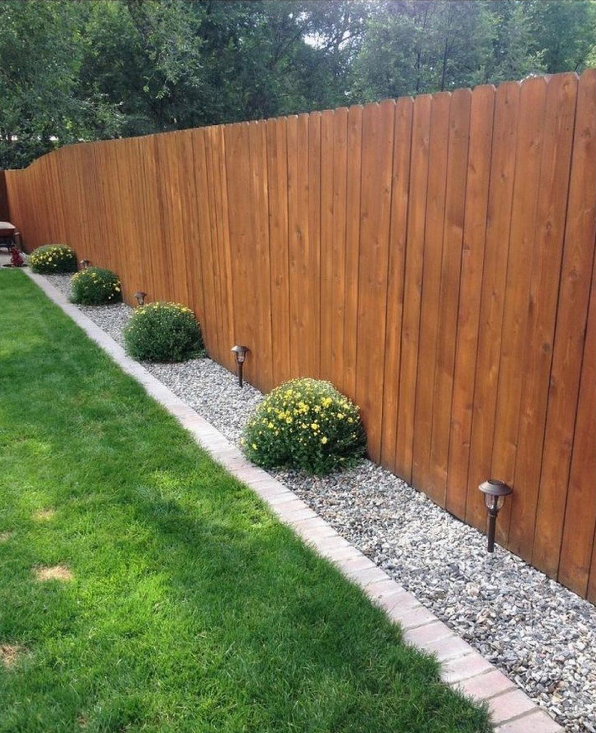 7 Backyard Fence Ideas for Privacy and Style 5 Tall vertical wooden privacy fence bordering a green lawn and gravel garden bed with shrubs.