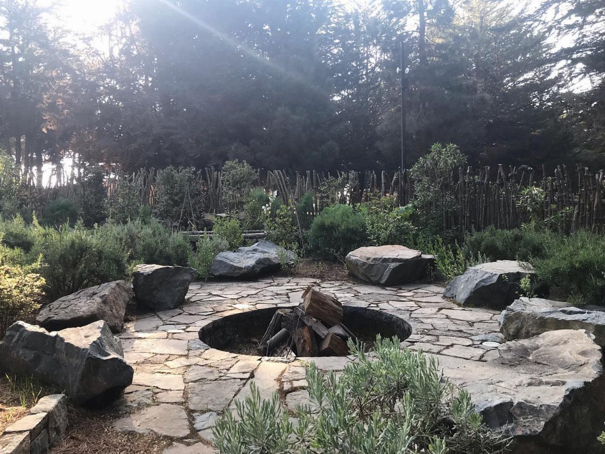 Stone fire pit with logs surrounded by large rocks and green garden plants.