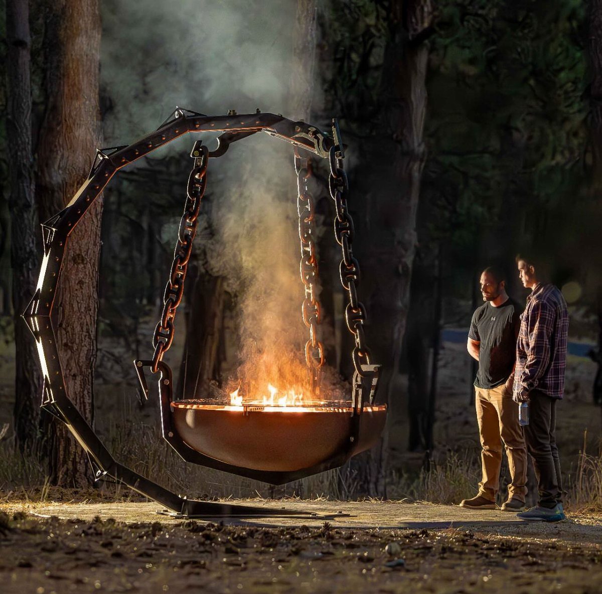 A large sculptural metal fire bowl suspended by chains from an arched frame in a forest.