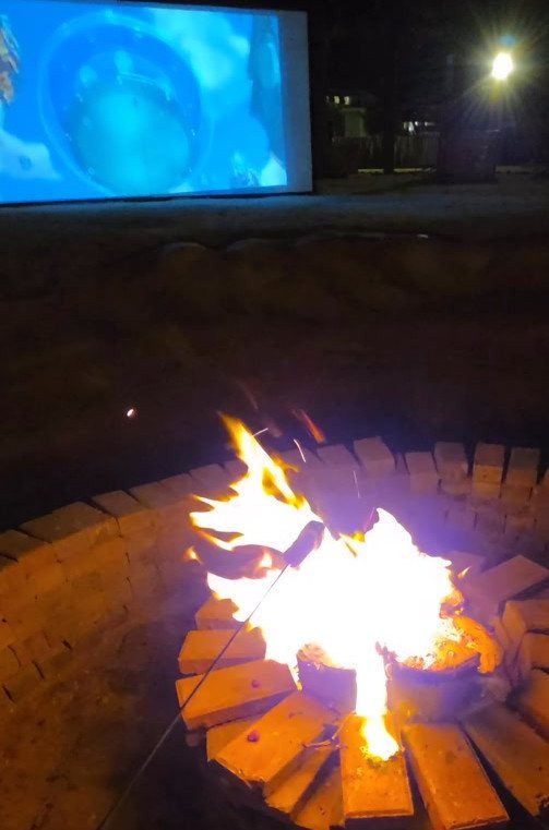 A glowing fire pit in the foreground with an outdoor movie screen in the background.