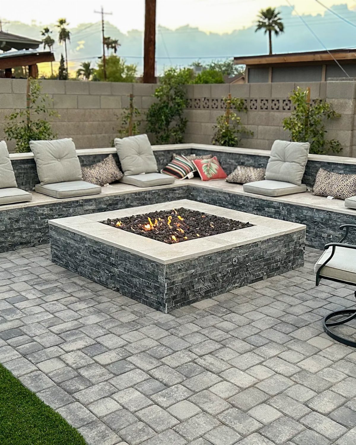 A square stone fire pit with glowing embers on a grey brick paver patio.