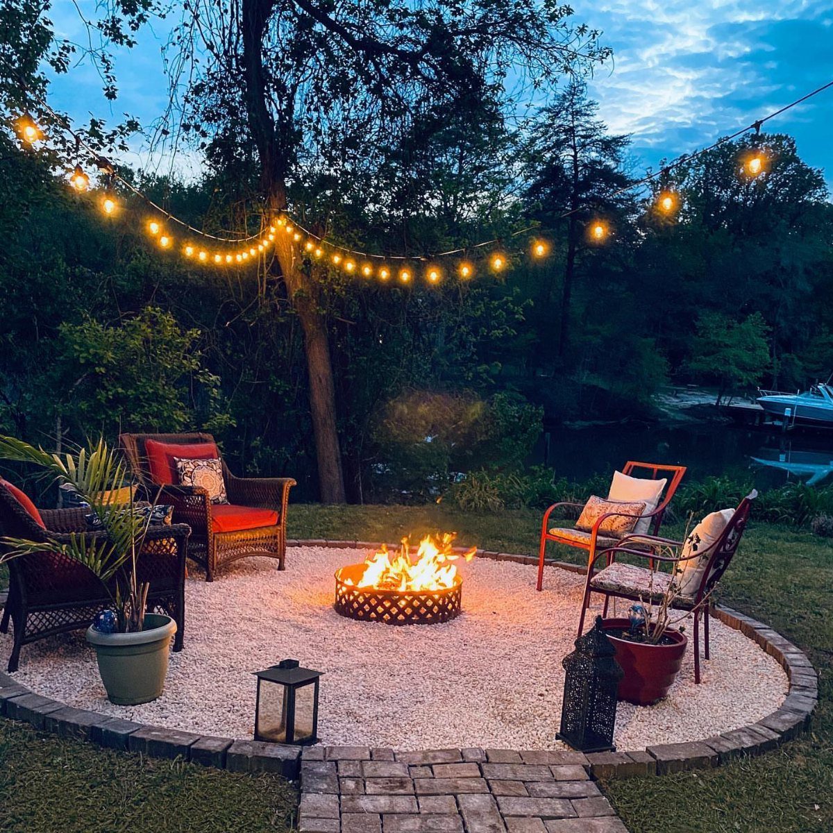 A circular gravel fire pit area with chairs and string lights at dusk.