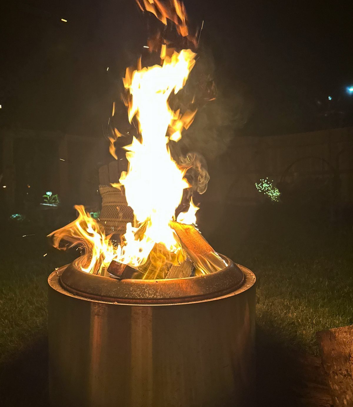 Bright orange flames rising from a cylindrical metal fire pit in a dark backyard setting.