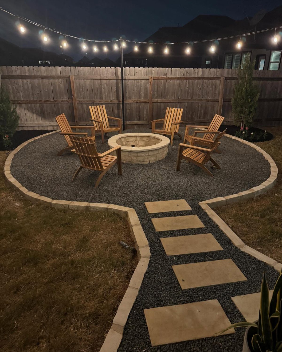A stone fire pit surrounded by wooden chairs on a gravel base with a stepping stone pathway.