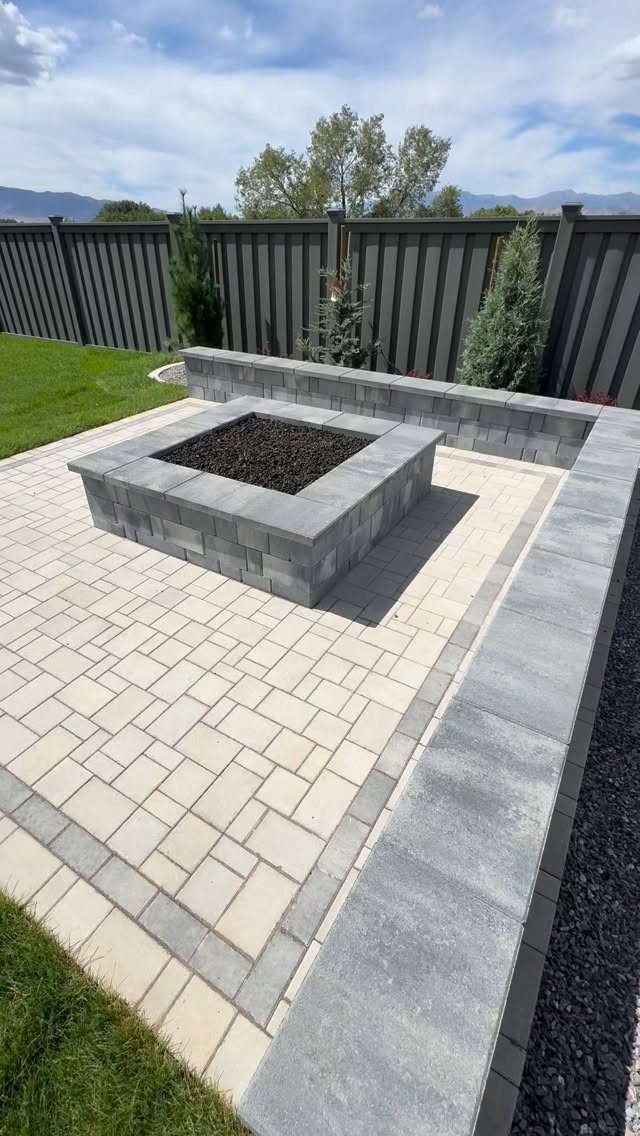 A square grey stone fire pit centered on a light paver patio with a low wall.