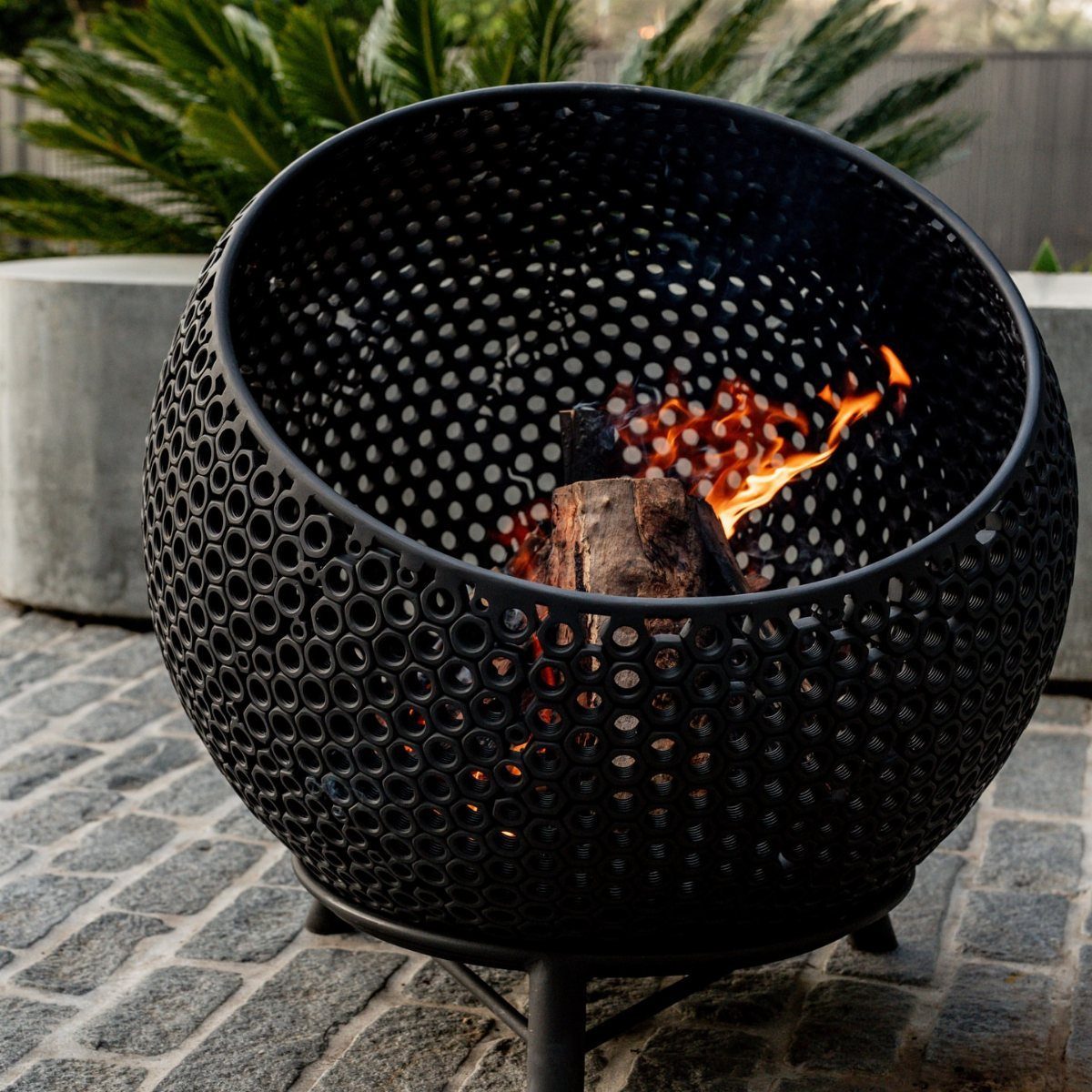 Black perforated metal fire bowl burning wood on a cobblestone patio.