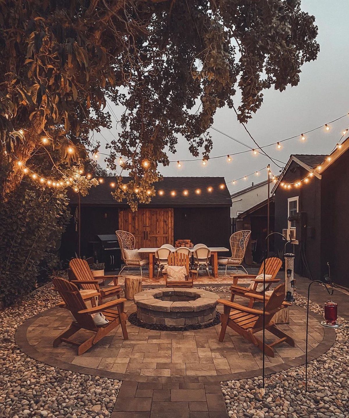A circular stone fire pit surrounded by wooden chairs on a paved patio under string lights.
