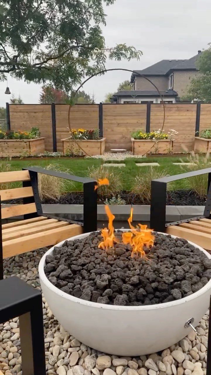 A white fire bowl with black lava rocks and orange flames sits on gravel.
