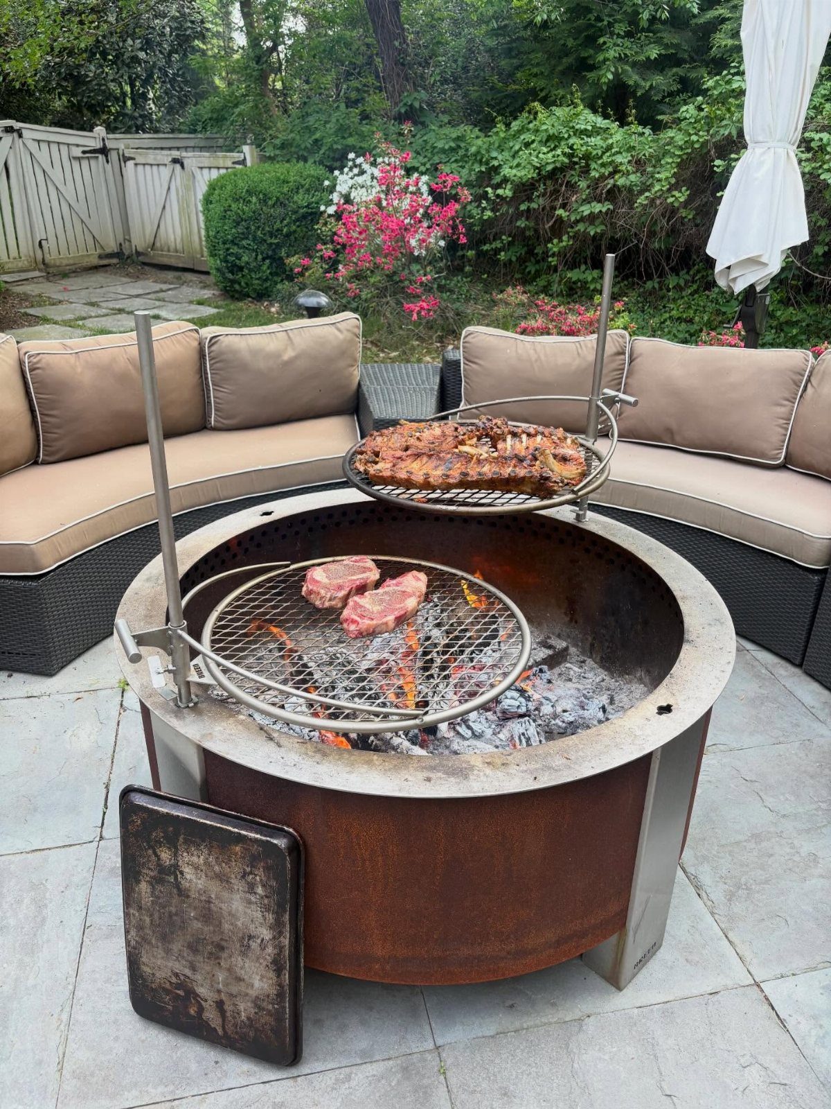 Metal fire pit with two cooking grates holding steaks over burning wood on a patio.