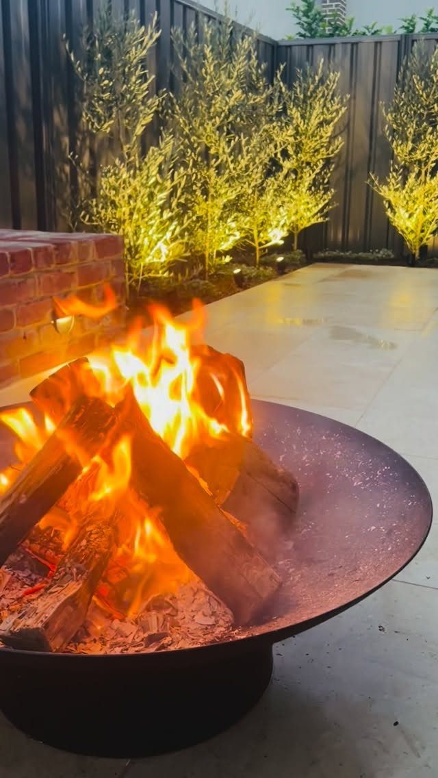 23 Backyard Garden Inspiration Ideas for Any Space 9 Burning logs in a black metal firepit on a paved patio at night.