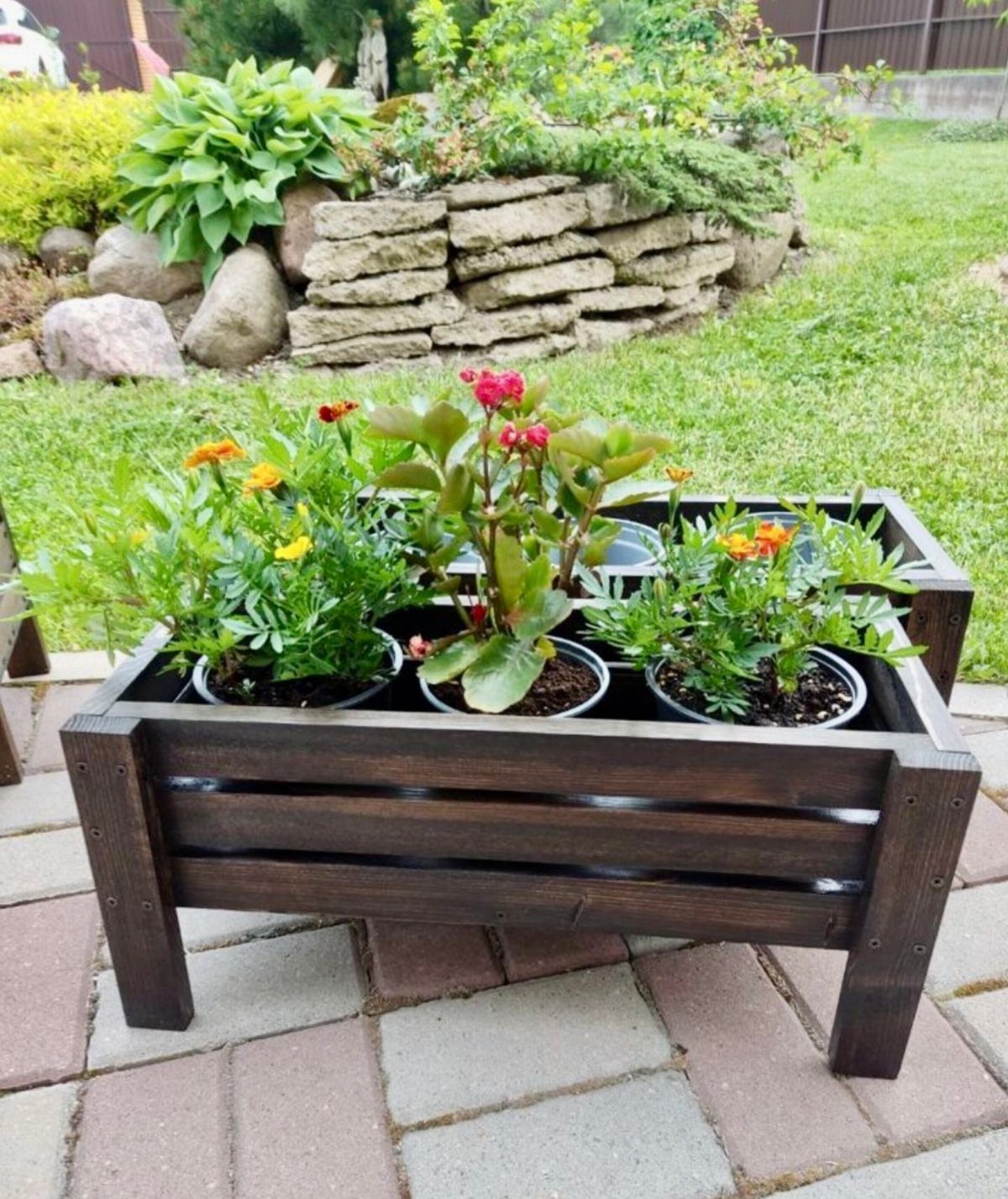 Dark stained wooden planter box filled with colorful flowering plants on a stone patio.
