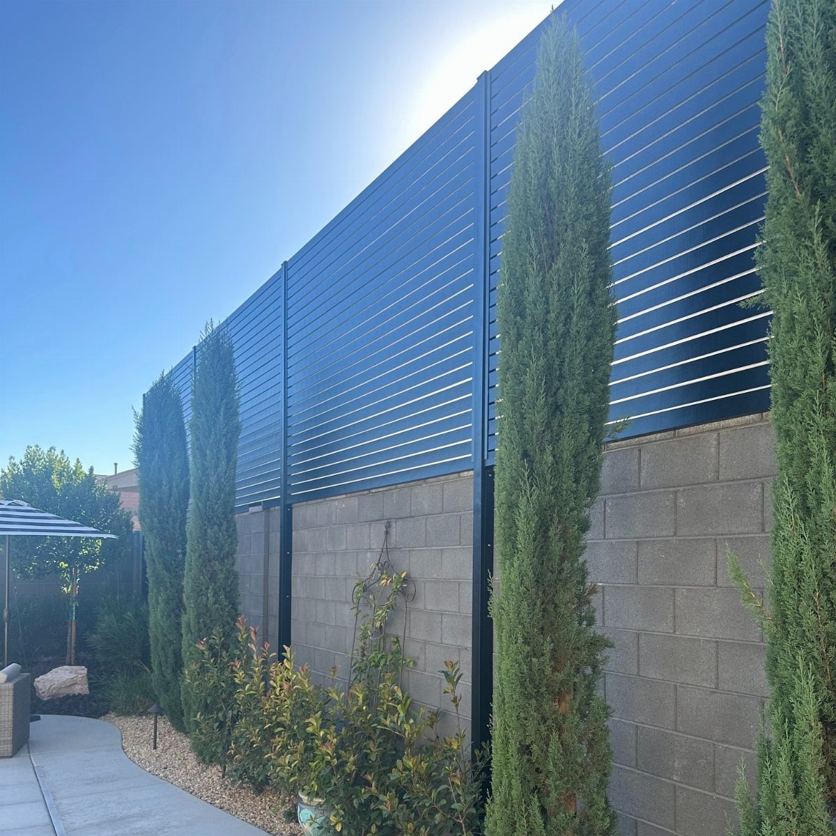 9 Backyard Patio Ideas with Fire Pits, Shade Sails & Retractable Awnings 4 Black horizontal slat privacy fencing installed atop a grey concrete block wall with tall evergreen trees.