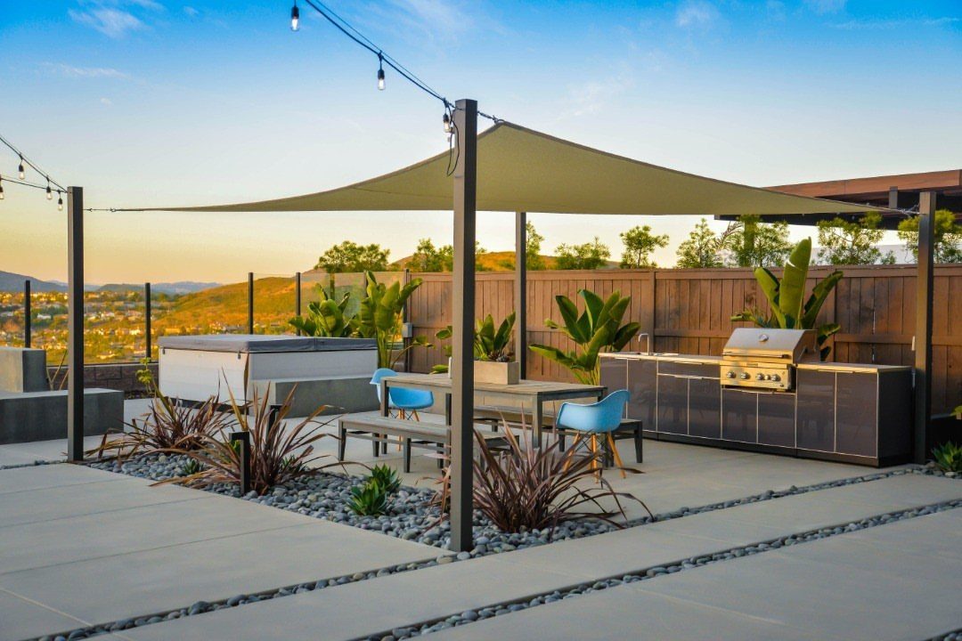9 Backyard Patio Ideas with Fire Pits, Shade Sails & Retractable Awnings 8 A large tan triangular shade sail covering a modern outdoor patio with dining furniture and kitchen.