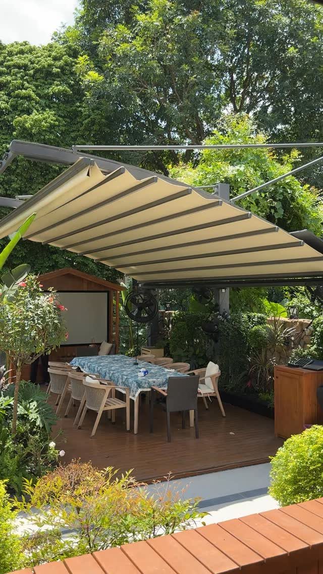 A beige retractable awning provides shade over a wooden patio dining area surrounded by greenery.