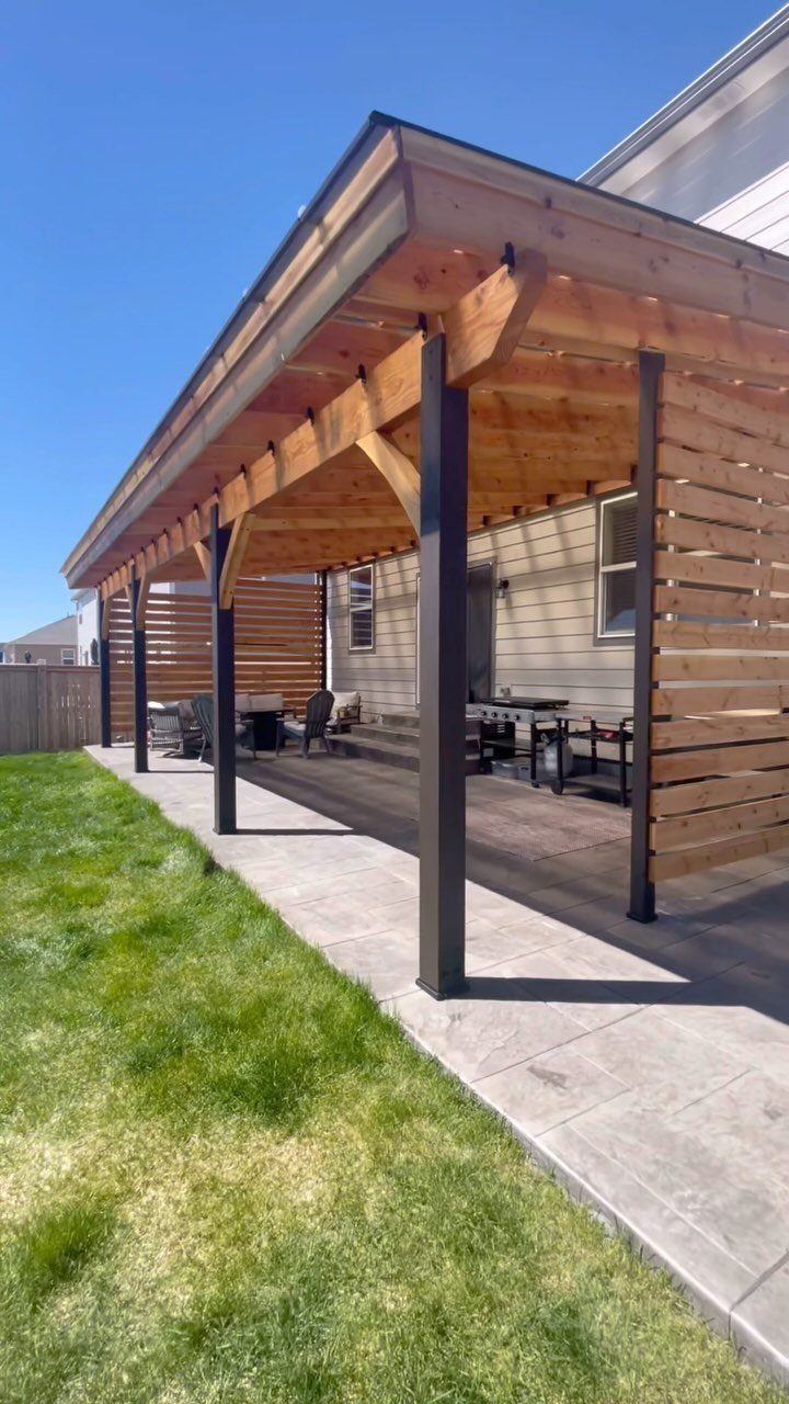 A wooden patio cover with dark metal posts shading a concrete outdoor living space.