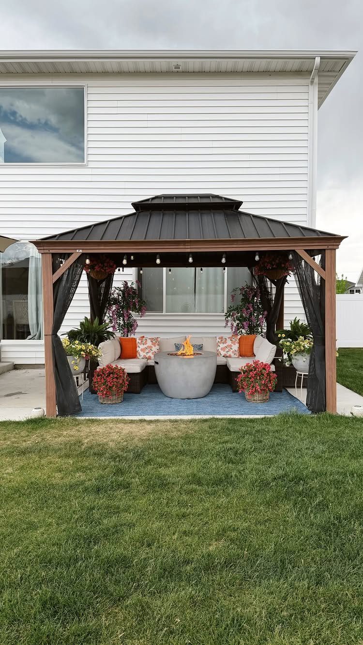 Wooden gazebo featuring white sofas and a central stone fire pit on a blue rug.