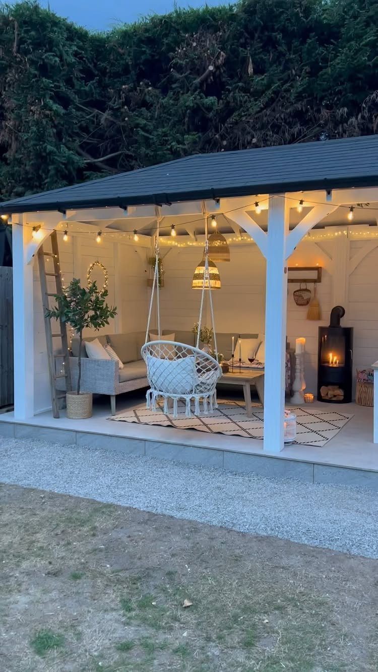 A white wooden gazebo containing a lounge area with a hanging chair and string lights.