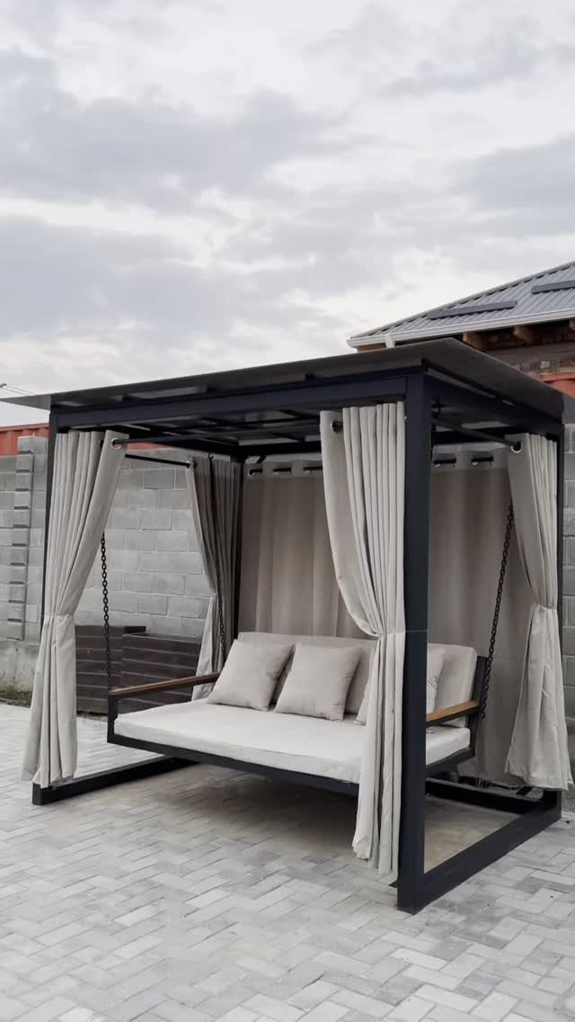 A modern black metal gazebo with a hanging beige daybed and flowing curtains on a paved patio.