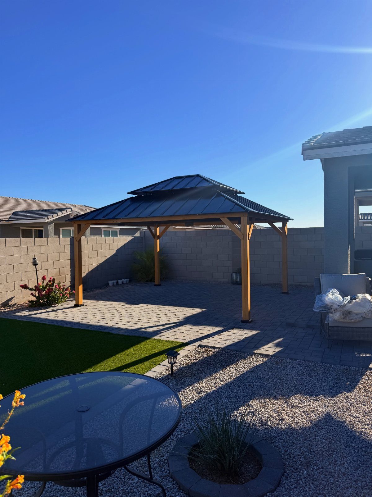 A wooden gazebo with a dark metal roof situated on a paved patio in a backyard.