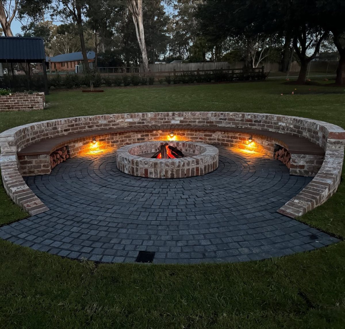 16 Beautiful Backyard Ideas for Cozy to Elegant Outdoor Spaces 3 A circular brick fire pit with a burning fire surrounded by a curved brick bench.