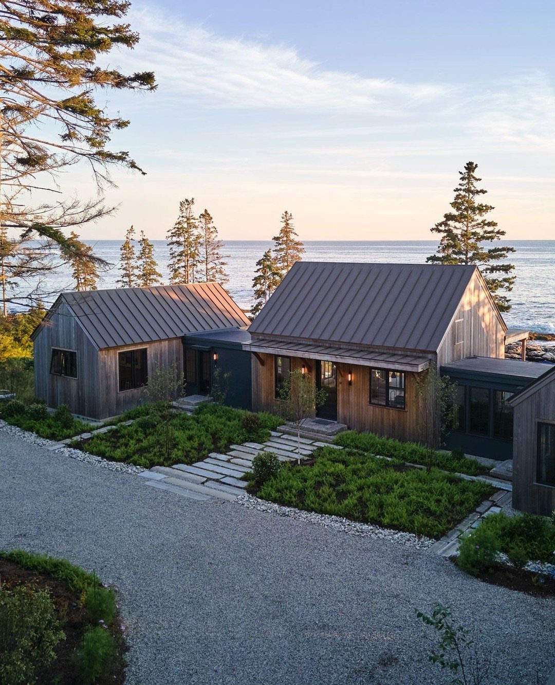 Modern wooden farmhouse buildings with metal roofs situated near the ocean at sunset.