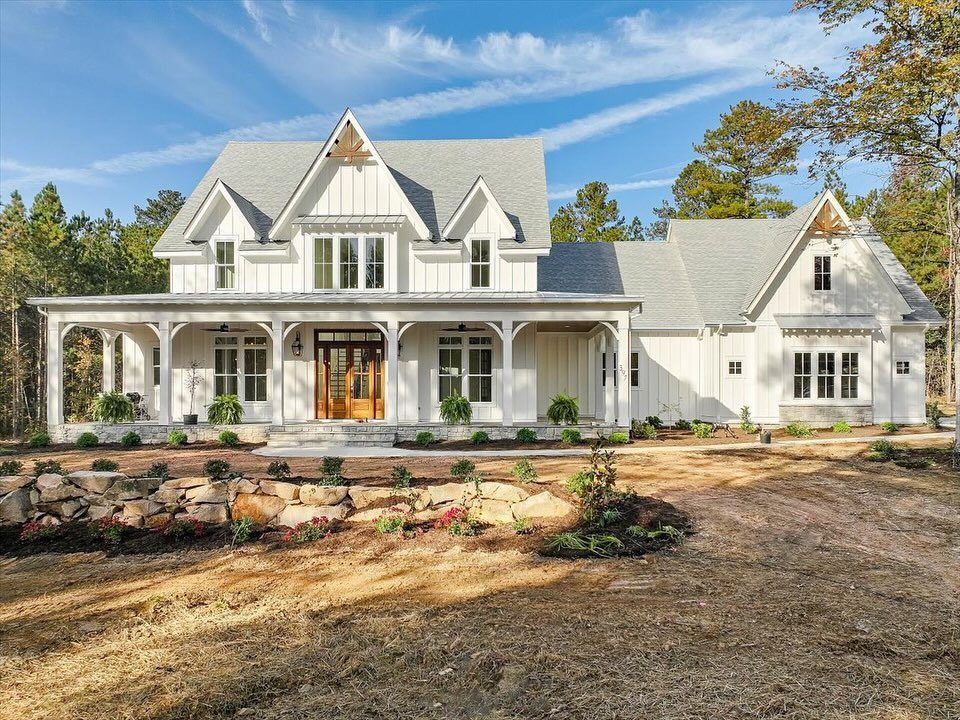 White modern farmhouse with a large wraparound porch and multiple gables under a blue sky.