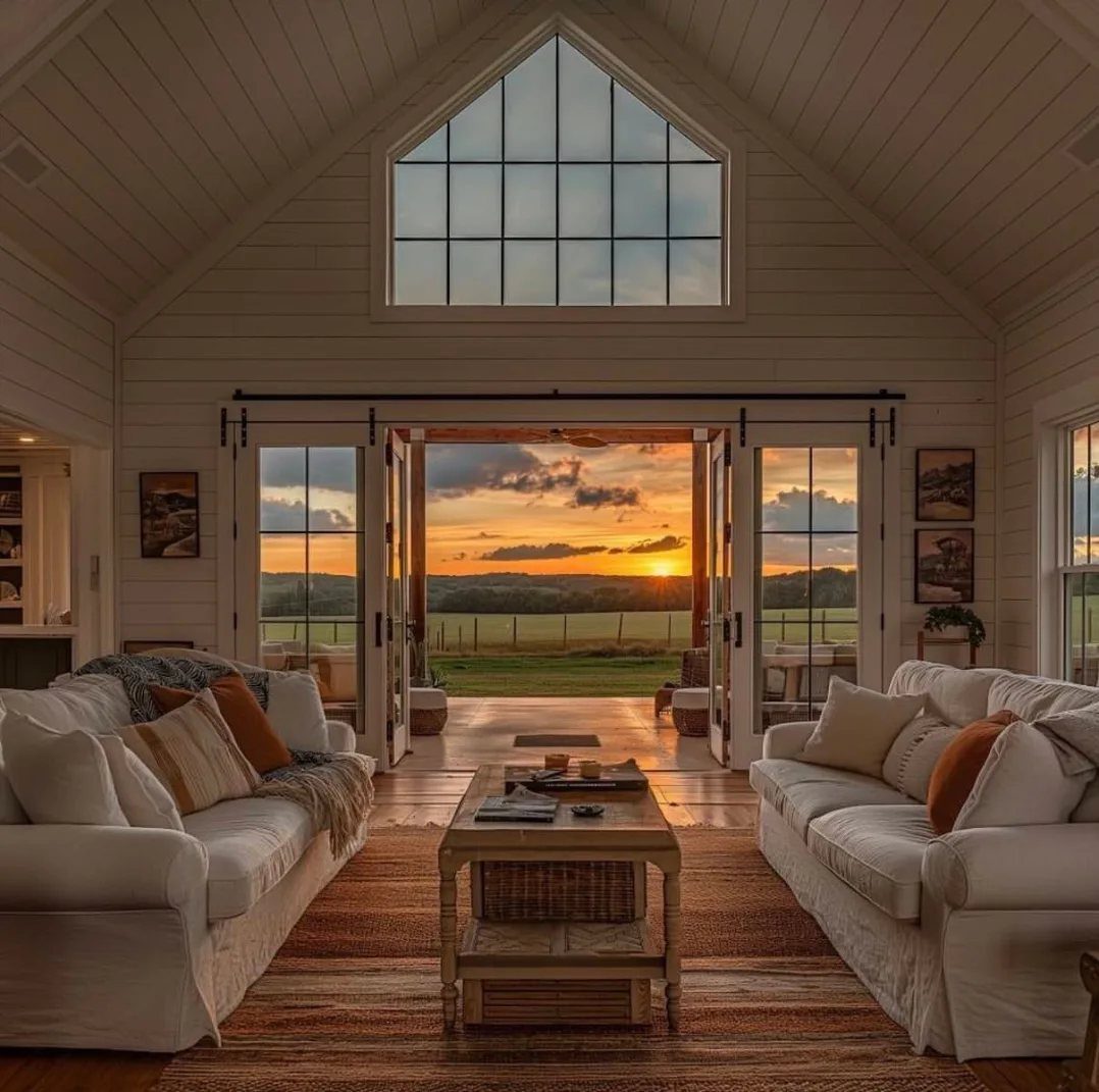 White sofas face large glass doors and a peaked window overlooking a sunset field.