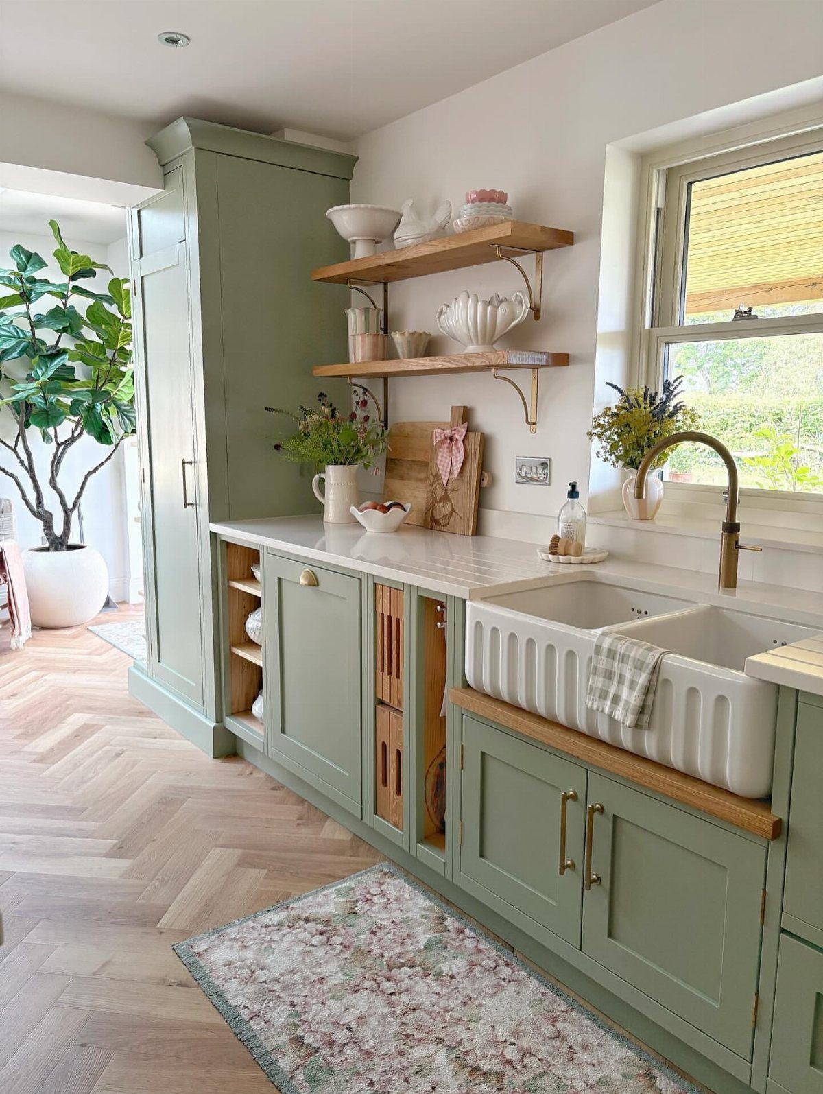 Sage green kitchen cabinets with a white farmhouse sink, wooden shelves, and marble countertops.