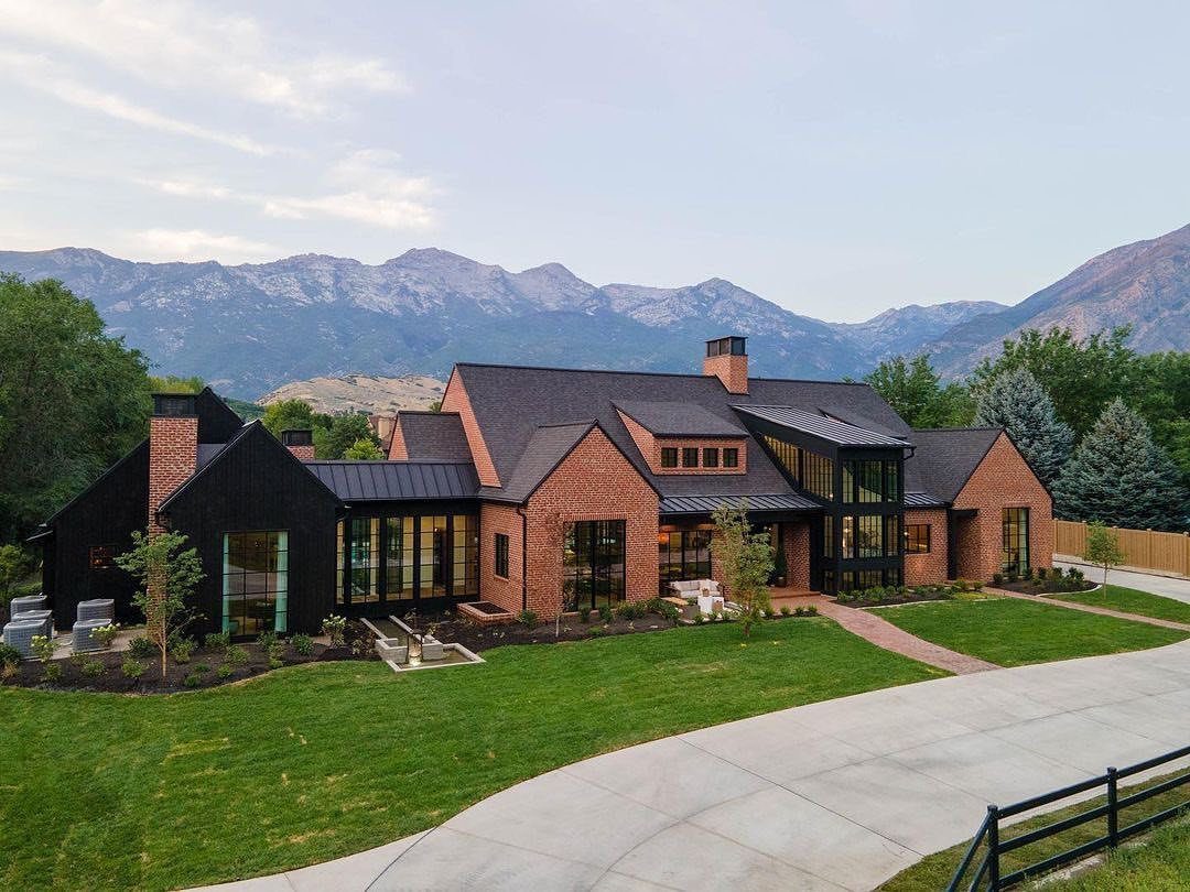16 Black Modern Farmhouse Exterior Ideas With Photos 13 A large modern farmhouse with black siding and red brick walls under a mountain range.