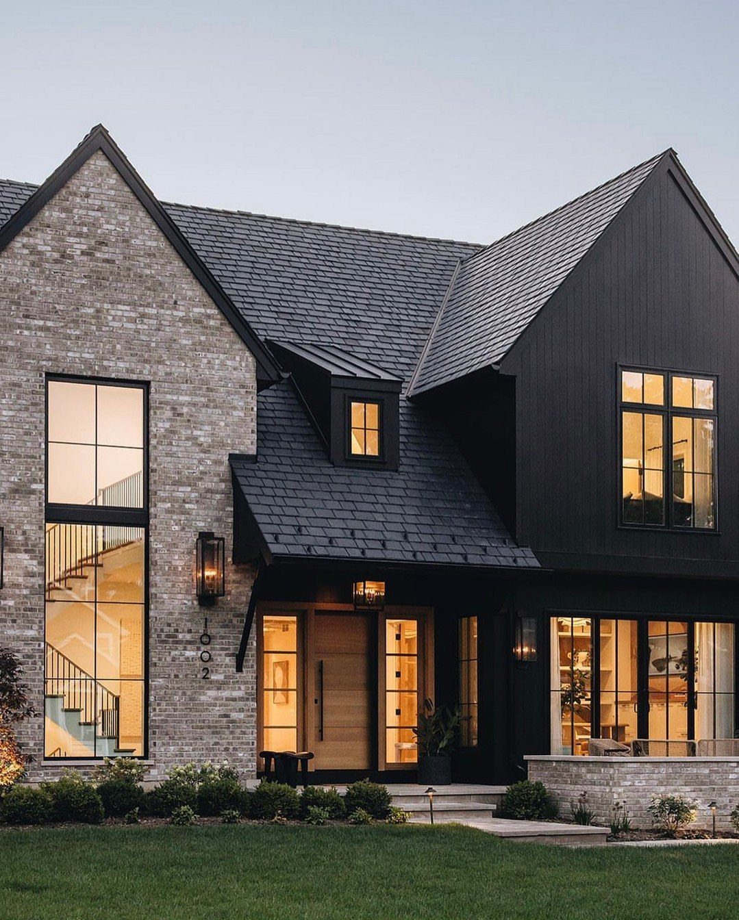16 Black Modern Farmhouse Exterior Ideas With Photos 14 A modern farmhouse featuring grey brick, black siding, large windows, and a wooden front door.