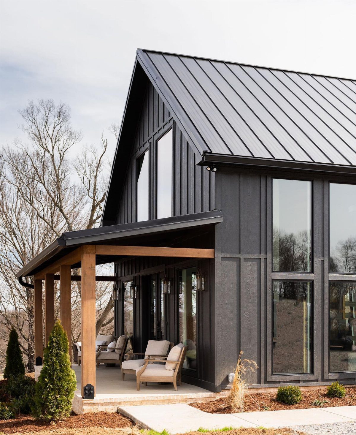 16 Black Modern Farmhouse Exterior Ideas With Photos 1 Black modern farmhouse with large windows, a metal roof, and a wooden porch area.