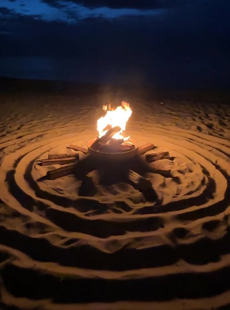 A bright bonfire burning in a metal ring on a sandy beach at night.