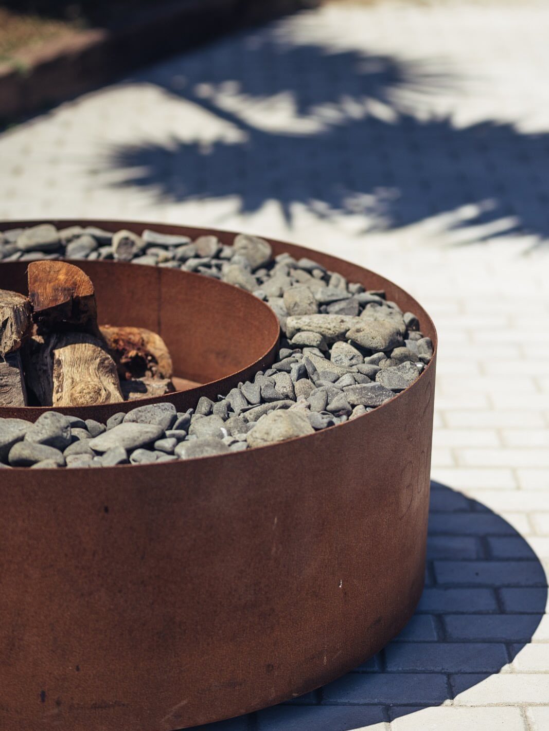 A circular rusted Corten steel fire pit filled with grey stones and wood logs.